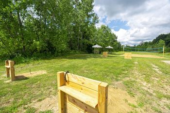 A wooden bench is in the foreground of a grassy area with trees and a volleyball net in the background.