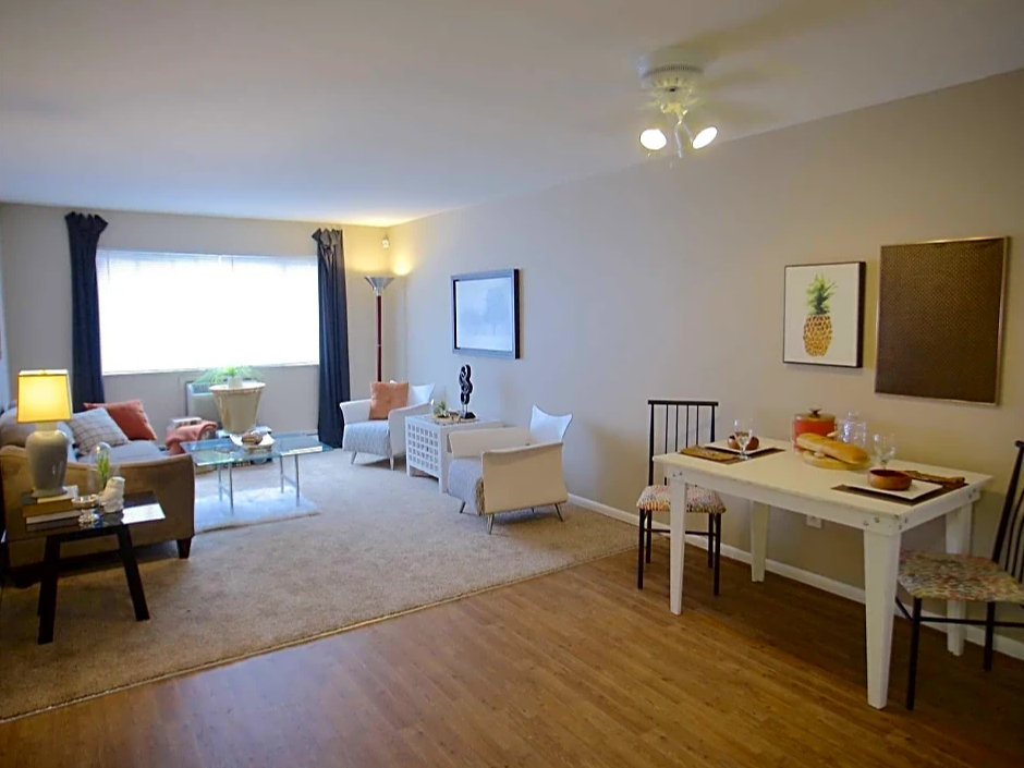 Photo taken from dining room with wood floor looking onto living area with beige carpet.  Dark curtains on back wall to the right and left of window.  Dark colored couch, light coffee table with dark end table.  Two light colored chairs on right wall with picture in the middle.