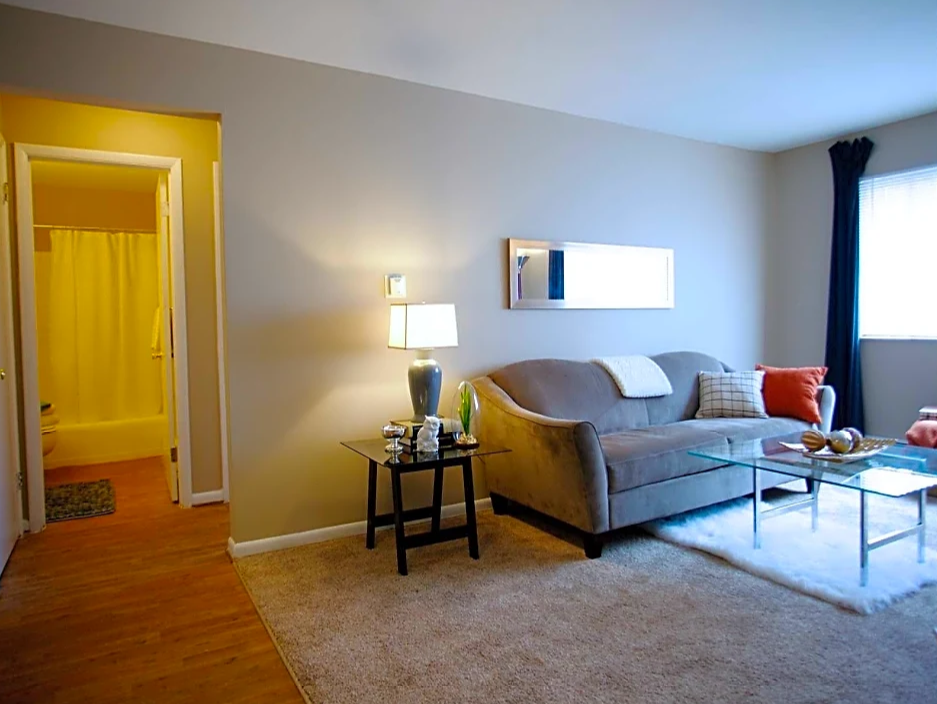 Living Room with brown couch, glass coffee table in front on beige carpet.  To the right on far wall is a window with dark curtains.  Room looks onto bathroom.