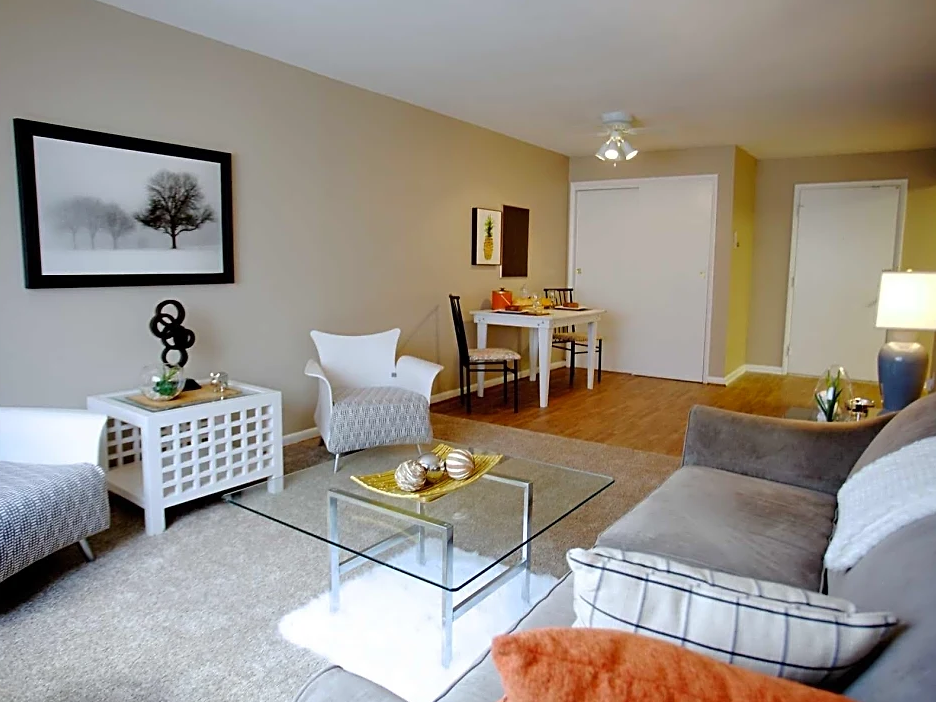 Living area with brown couch on right wall with pattern pillows, glass coffee table in front, with two white chairs and end table on left wall.  Artwork above table in center.  In background is dining area and lamp.