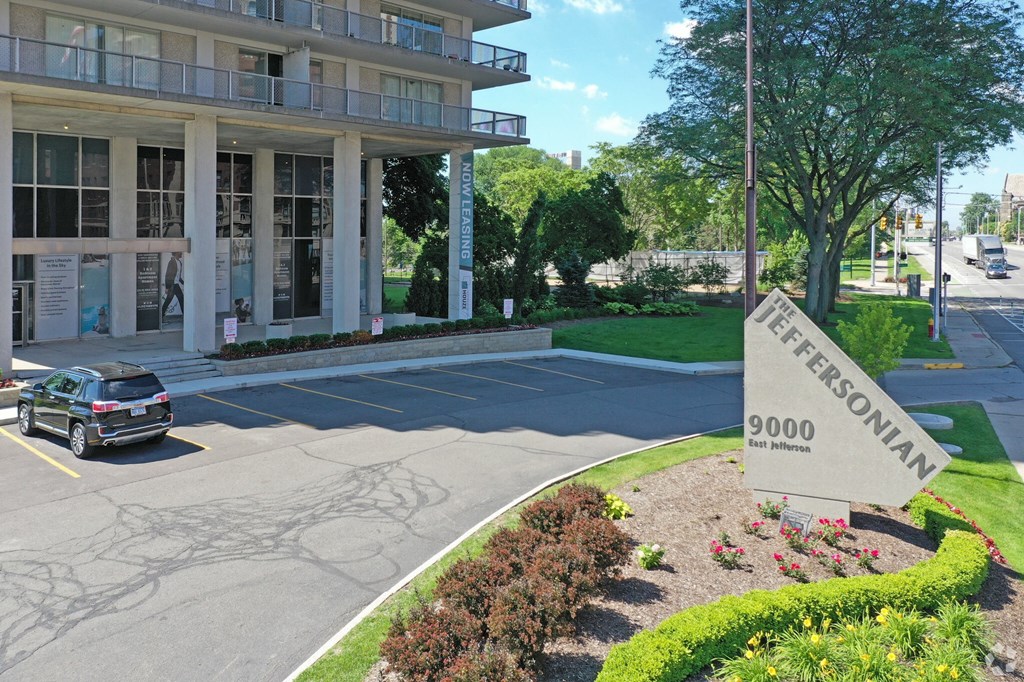 Outdoor image of the Jefferson. The parking lot is empty with the Jefferson sign next to the street. There are green bushes and blue skies.
