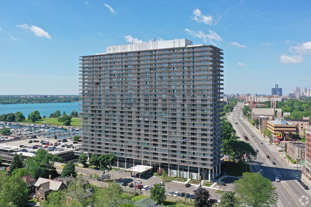 Exterior of The Jefferson. The building stands 30 stories tall. The building is made of concrete and glass. There are green trees and blue skies. You can see the Detroit River in the background. The building is located on Jefferson Ave.
