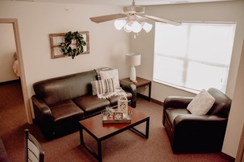 Interior of the living room in an apartment with brown couch, chair, white ceiling fan and coffee table. Back right has window. - Photo Gallery 2