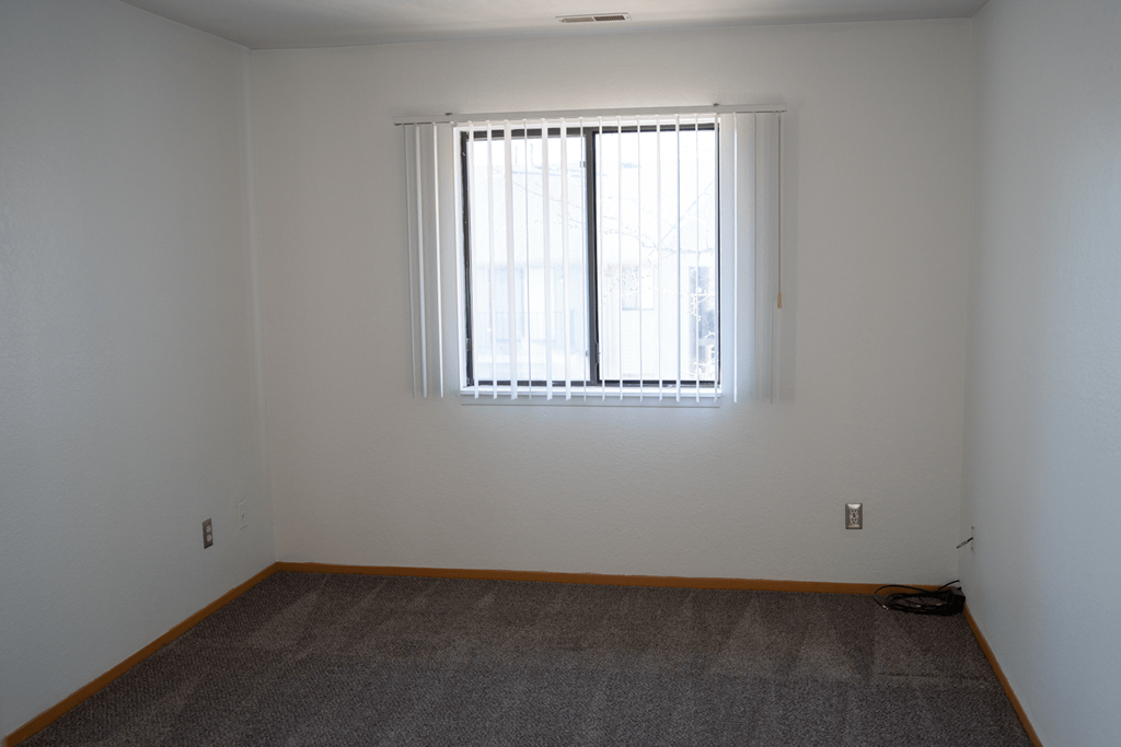 Bedroom at Glenwood with plush grey carpeting, white walls, and brown trim. In the center of the room is a large window with blinds that are open allowing the sunlight to come into the room.