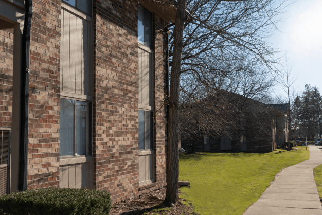 Exterior of Glenwood Apartments. The building is made up of brown brick and brown siding. There is a tree and bush in front of the building. In front of the landscaping is a sidewalk that leads to another building. In between the two buildings is a large grassy area.