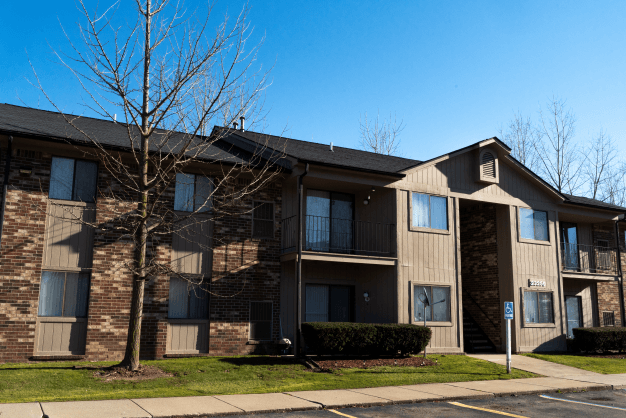 Exterior of Glenwood Apartments. It is a two-story brown building with patios and balconies. The sky is blue above the building. In front of the building is green grass, a sidewalk, and a parking lot. In front of the building to the left is a tree with no leaves.
