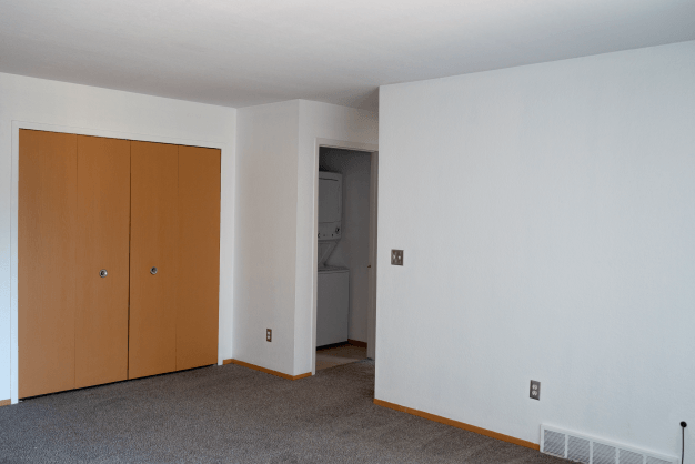 Living room at Glenwood with white walls, brown trim, and plush grey carpeting. To the left of the photo are two brown bifold closet doors. There is an opening leading to the hallway. A closet is open showing a white stacked washer and dryer.