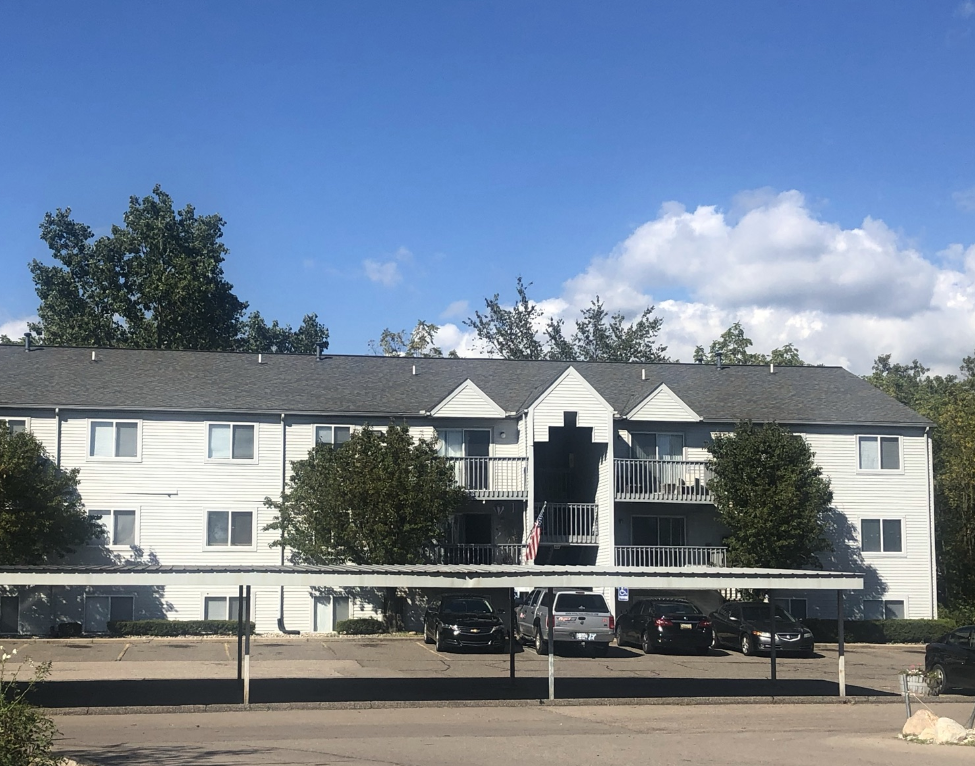 Drive into Greyberry Apartments with the building in the background.  The building is three stories and is grey in color with a dark grey roof. In between the drive and the apartment building is a grey carport. Behind the building are large green trees.