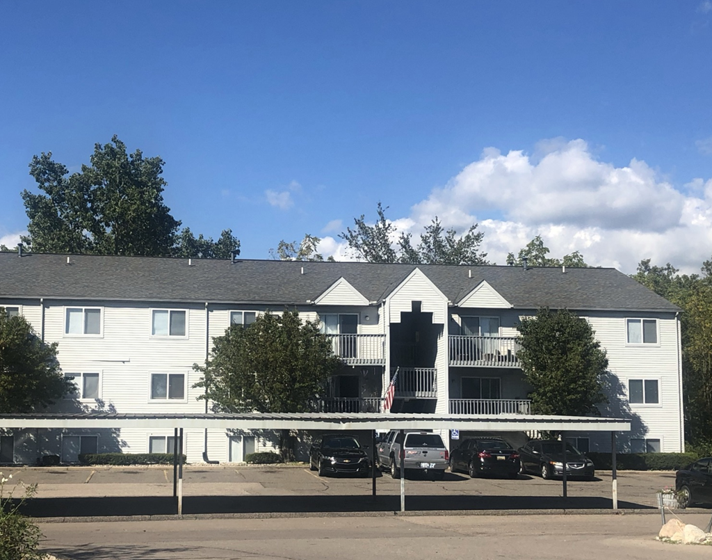 Drive into Greyberry Apartments with the building in the background.  The building is three stories and is grey in color with a dark grey roof. In between the drive and the apartment building is a grey carport. Behind the building are large green trees.