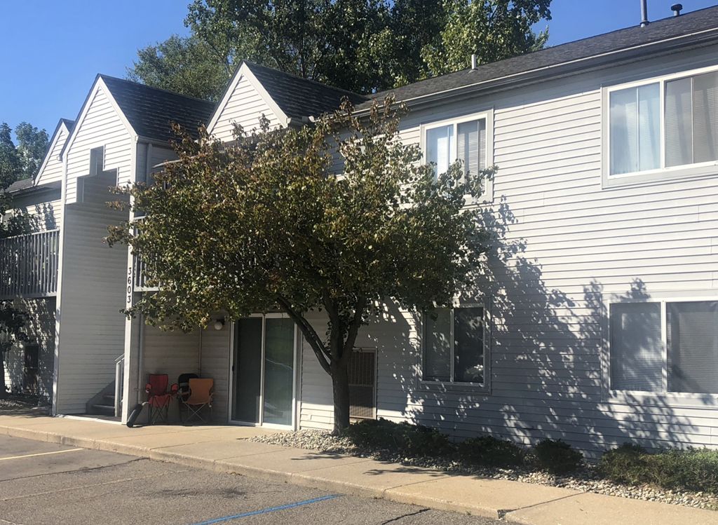 Two story grey apartment building with sidewalks leading up to the building.  There is a patio to the right of the breezeway. In front of the building is a tree and other green bushes.