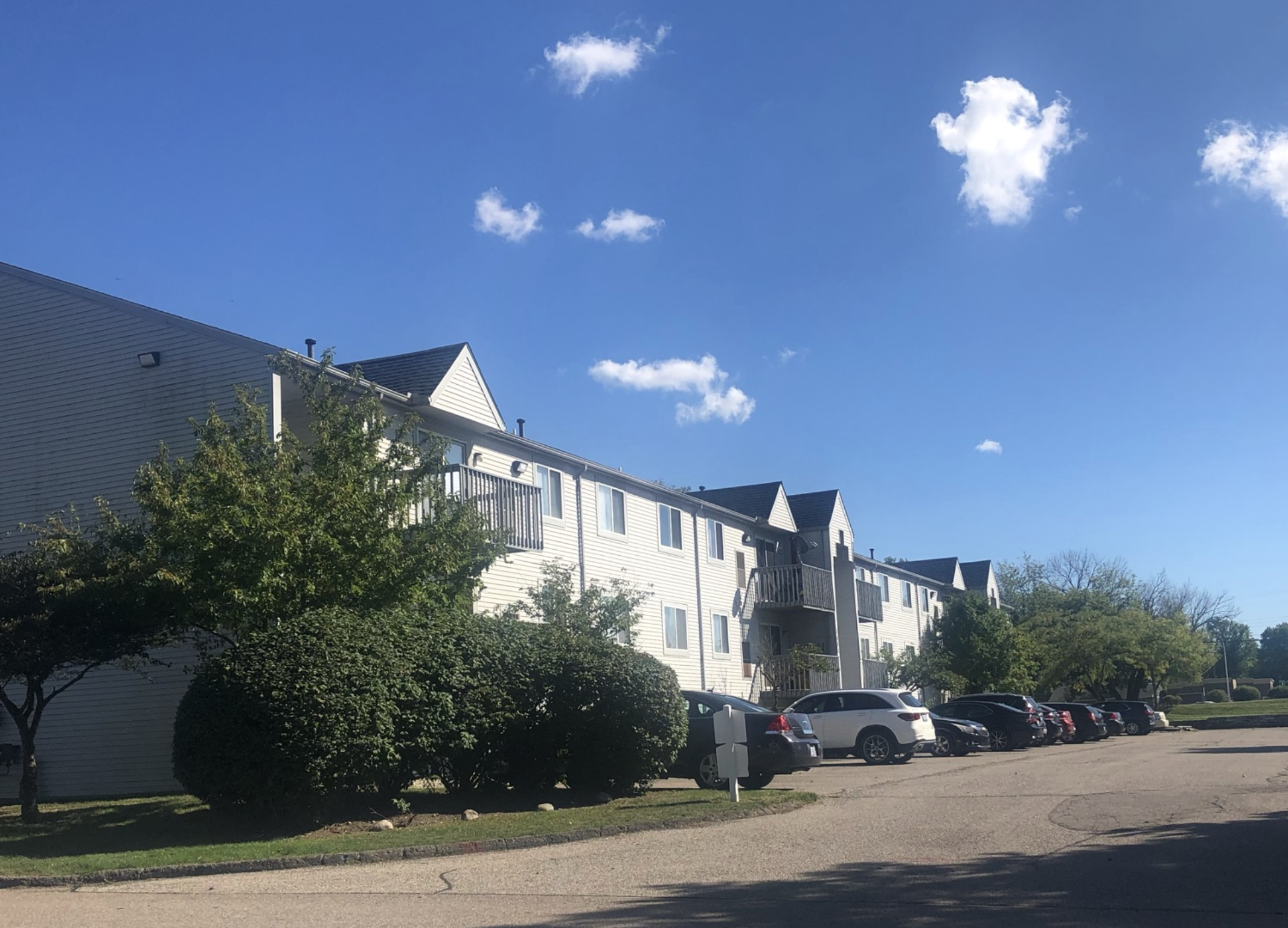 Three story exterior of a grey apartment building.  There are balconies on the third floor of the building and lush green landscaping.  The building in to the left and to the right is a grey carport with cars parked under it.