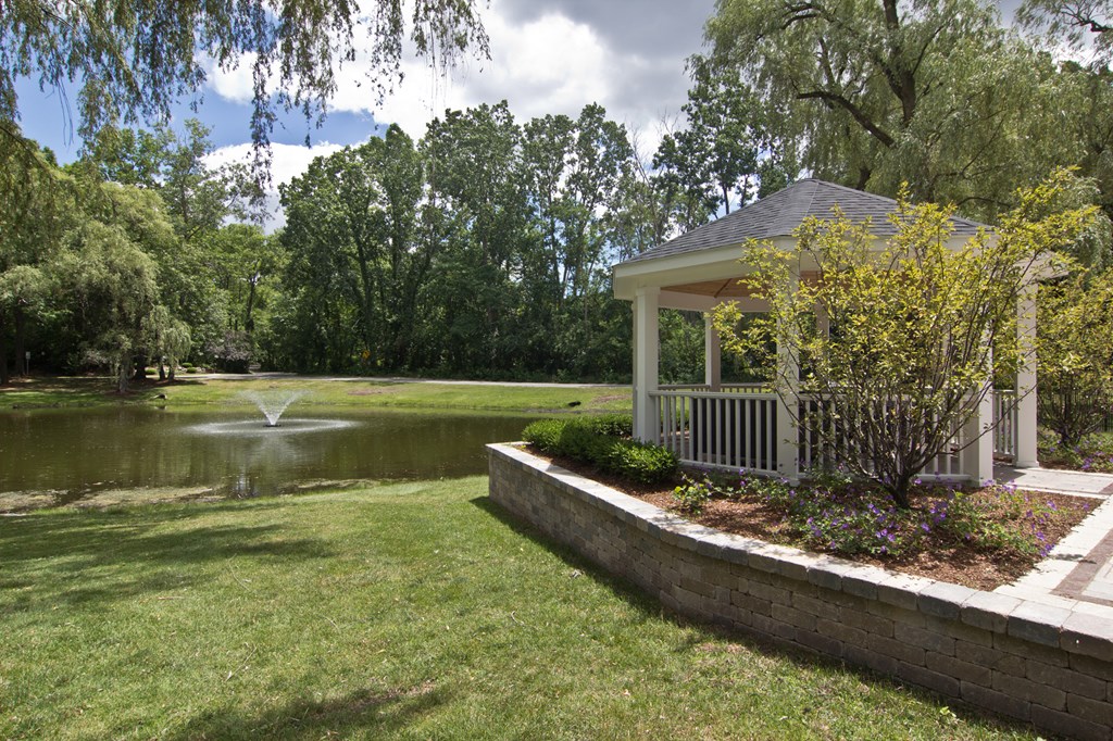 Gazebo And Fountain View White gazebo to the right of photo with trees, greenery and lined with wood surround. Green grass in the middle leading up to a pond with a fountain. Sky and green trees in the background.