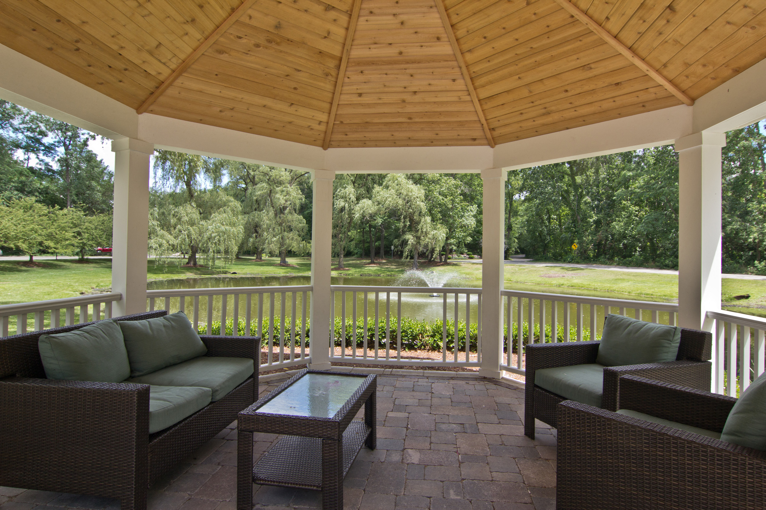 Interior of gazebo with with light wood ceiling and white trim. Wood slatted surround with brick flooring.  Green couch with coffee table to the left, and two green/brown chairs to the right.