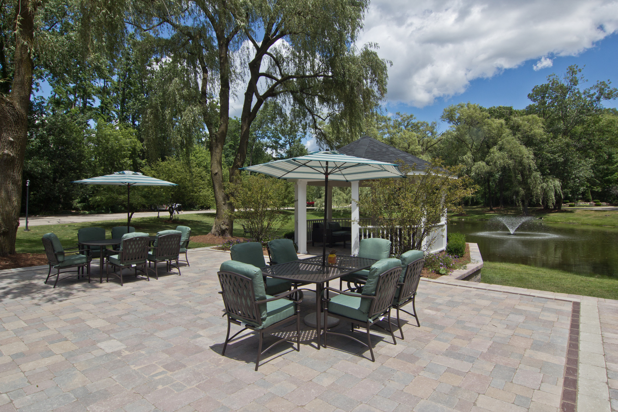 Patio furniture on brick decking overlooking the gazebo and pond with fountain.  Trees and blue sky in background.  2 Tables all have 4 green chairs and umbrella.