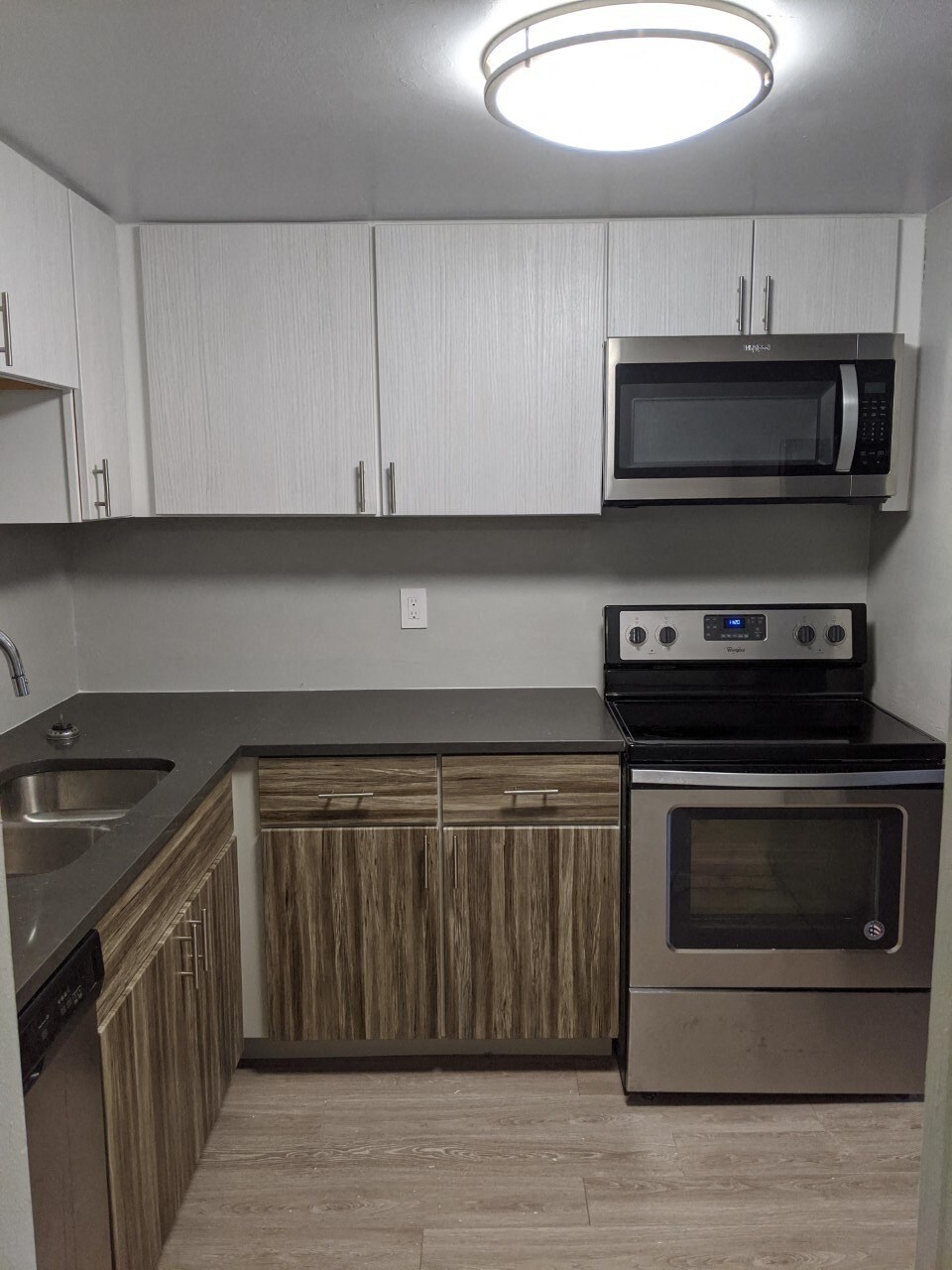 Image of kitchen, there are dark wood-like cabinets below with white cabinets on top. There are black countertops and stainless steel appliances. There is a sink to the left and an oven with a microwave above on the right. The walls are a light grey.