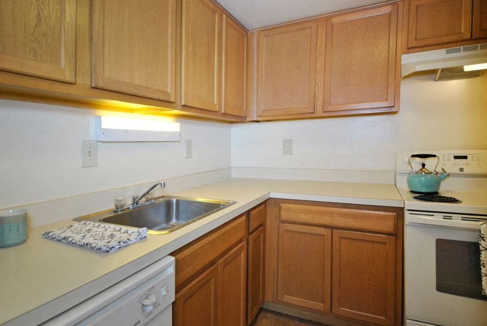Photo of a kitchen, there are light wood cabinets below and above. The countertops ate white followed with white appliances. There is a sink and dishwasher to the left and a stove on the right. The walls are white and there are lights above the sink and oven.
