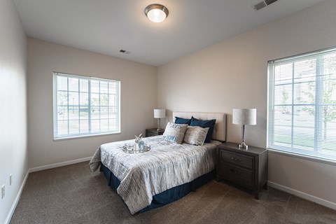 Bedroom, large bed in center of room with matching end tables on each side, window to the left and to the right of the bed, light tan painted walls with tan carpet.