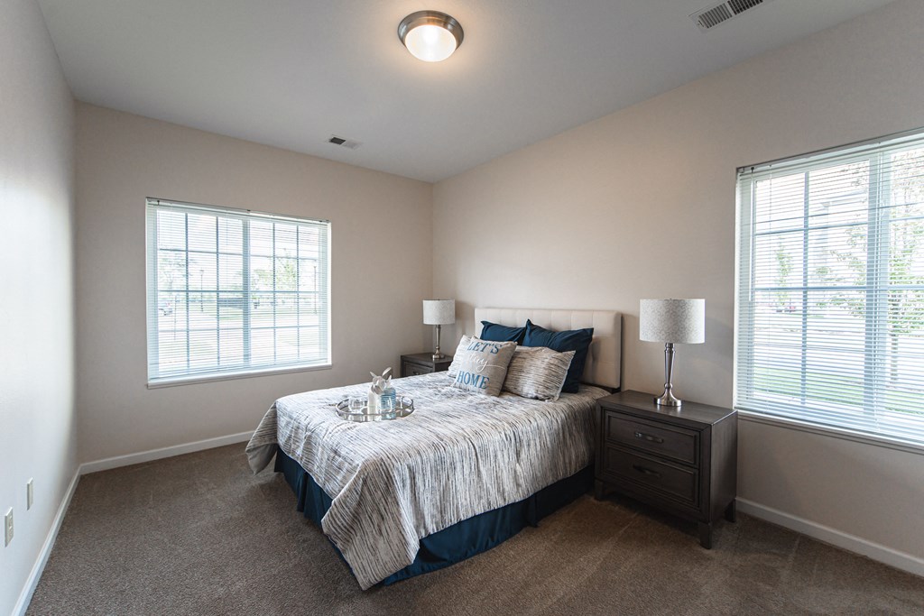 Bedroom, large bed in center of room with matching end tables on each side, window to the left and to the right of the bed, light tan painted walls with tan carpet.