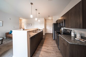 Entrence into kicten, dark brown cabinets with light grey granite countertops, island to the left, stainless steel black appliances, white walls.