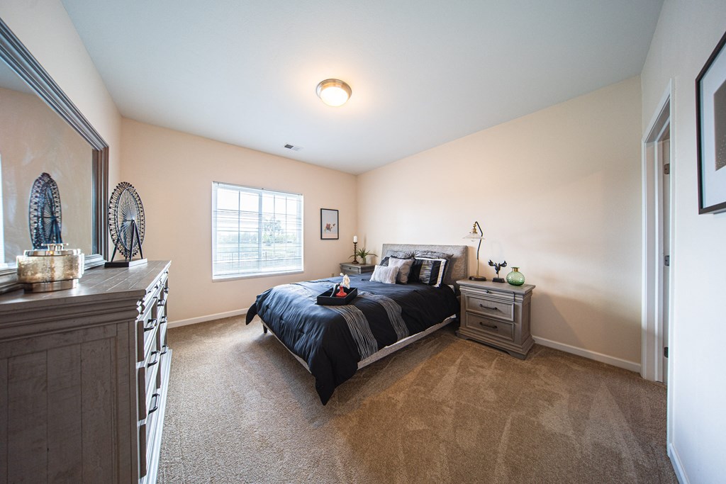 Bedroom with large bed in the middle of the room, window on left side of room, matching end tables on each side of bed, dresser across from bed in room, light tan walls, tan carpet.