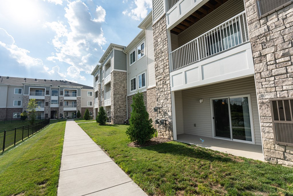 Paved sidewalk along the community, green grass, patios of apartment homes on right, fence next to pond on left.