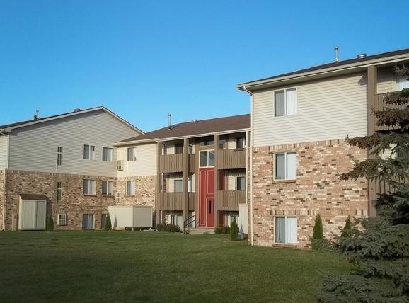 Exterior of Brighton Cove on a bright sunny day. There is an expansive green lawn in front with a large pine tree to the right of the photo. The building is three forths brown brick from the bottom up and then brown siding for the reminder of the building. The building is 3 stories and there are brown wooden balconies on the second and third floors. The sky is bright and blue.