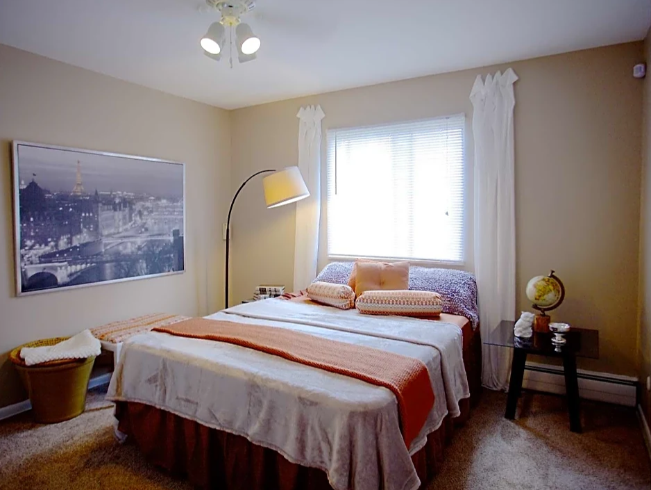 Bed against window with white curtains on each side of window.  Two-tone beige walls with white accent trim.  Striped orange bedding, beige carpet and photo on left wall.  Basket under photo and table to right of bedside.