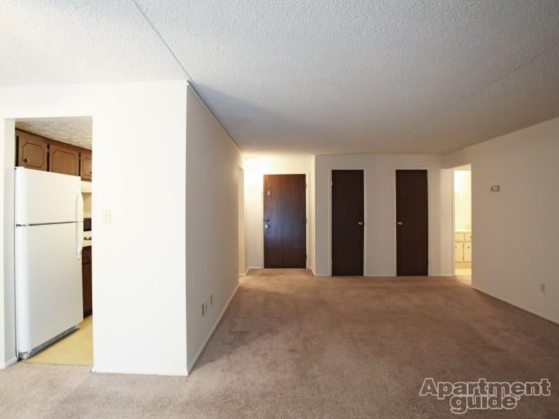 Living area with beige carpeting, dark brown doors to coat closet and exit of apartment, looking onto kitchen