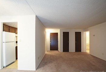 Living area with beige carpeting, dark brown doors to coat closet and exit of apartment, looking onto kitchen - Photo Gallery 12