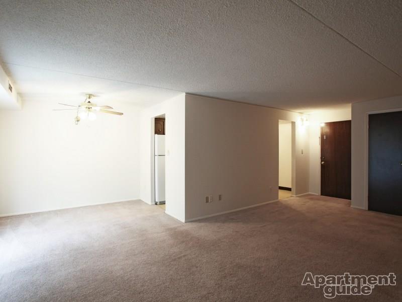 Living area with beige carpeting, ceiling fan, and doorway leading to galley style kitchen, door to right leading outside of apartment.