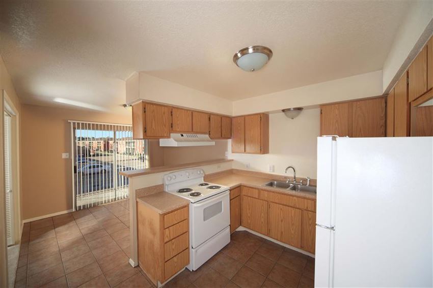 Kitchen with white appliances, stainless steal sink, brown cabinets, and tile like floor. The kitchen is open to the dinning room that opens to the patio with sliding glass doors. - Photo Gallery 1