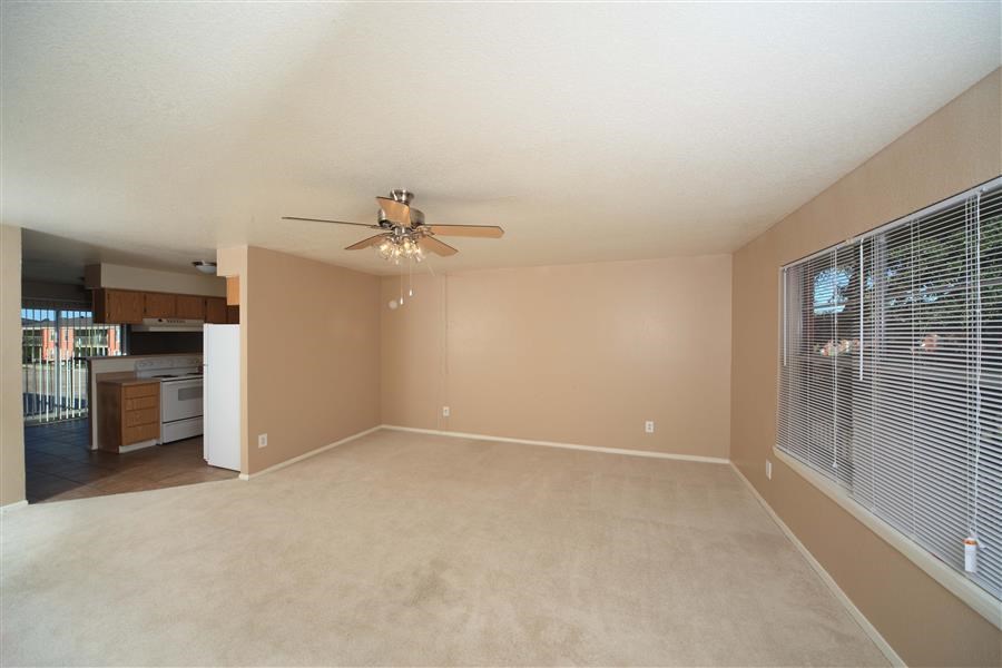 Living room with a large picture window to the right, and the kitchen to the left. The living room has brown walls, brown plush carpeting, and a ceiling fan.