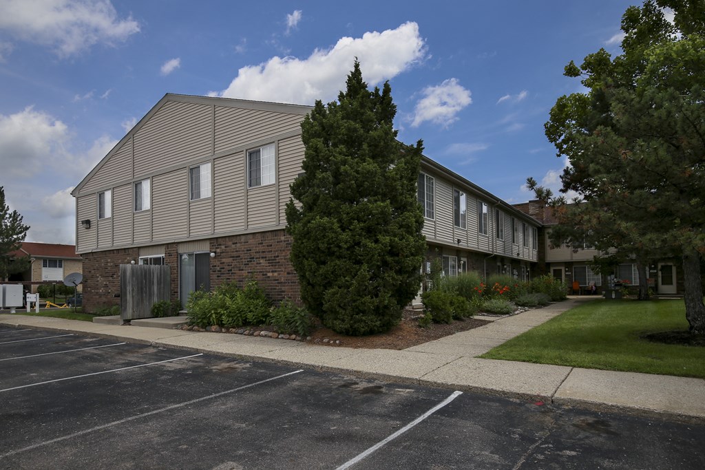 an apartment building in a parking lot with a sidewalk and a tree at Village Club of Rochester Hills, Michigan