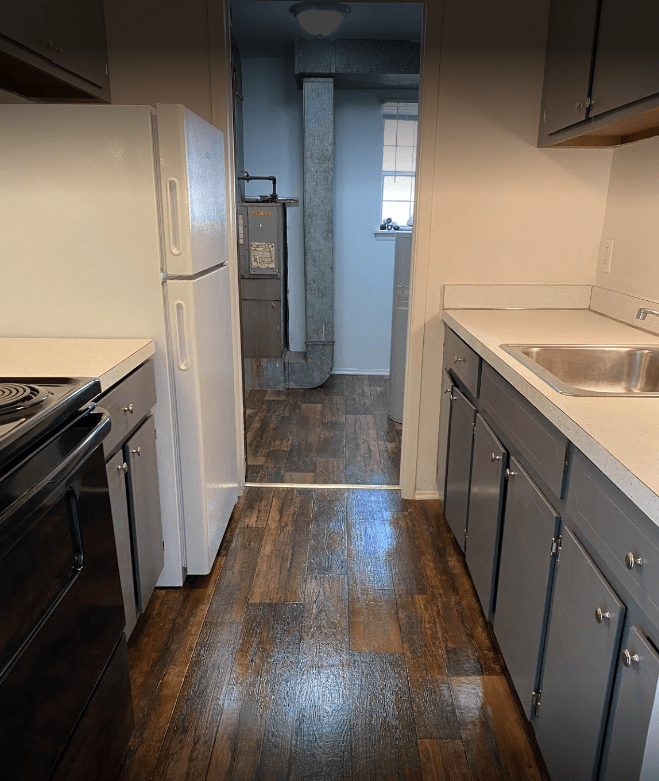 Galley style kitchen with dark cabinets, and dark wood flooring.  White countertops and black stove, white fridge.  Utility room off back of kitchen.