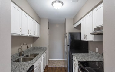 Galley-style  kitchen with granite speckled grey/white counters, white cabinets with pull handles on both lower and upper, hardwood-like flooring and stainless appliances