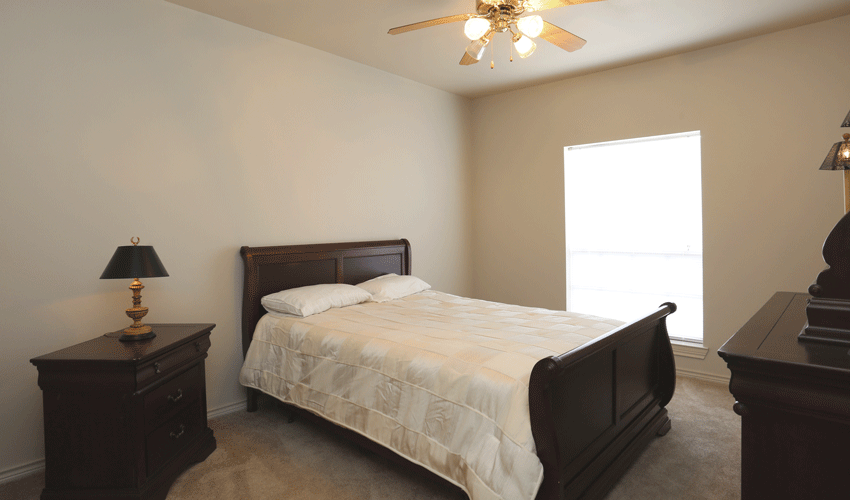 Bedroom with tan carpeting and tan walls. There is a large window and ceiling fan in the bedroom. There is a dark brown sleigh style bed with light bedding.