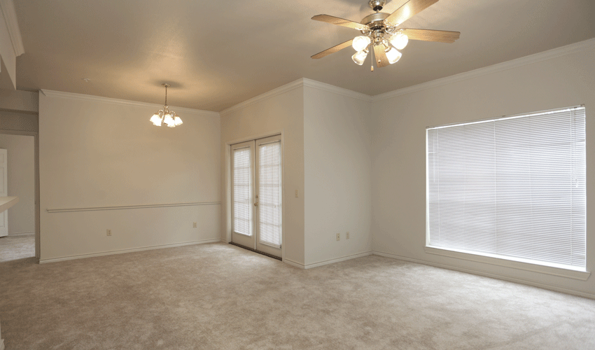Living room and dining room with tan carpeting and tan walls. In the living room there is a white ceiling fan and in the dining room there is a chandler.