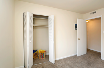 Bedroom with closet open. The closet has two white bi-fold doors. The bedroom has tan carpeting and cream colored walls. To the right is an open white door leading to the hallway. - Photo Gallery 12