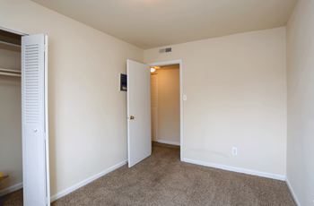 Bedroom with plush tan carpeting and cream colored walls. To the left there is a bi-folded white closet door open. - Photo Gallery 11