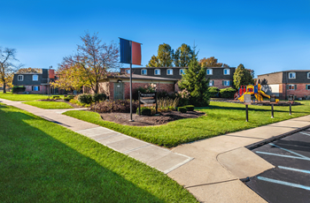 Exterior courtyard and parking lot. Sidewalk leading to leasing office. In the middles of the photo is a courtyard and playscape. Past that is the exteriror of Winchester Village. To the right of the photo is a black asphalt parking lot.