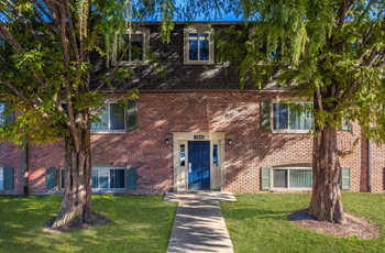 Exterior of Winchester Village Sidewalk leading to entryway of brown three story brick building with blue door. There are two large trees on either side of the blue door.