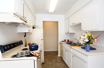 Galley style kitchen with brown tile flooring, white cabinets and appliances, and white counteriops. In the back of the ktichen is a closet/pantry.  There is a boquet of flowers in a blue vase on the counter ot the right. - Photo Gallery 10