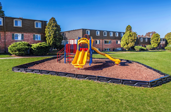 Playscape Red, yellow and blue colored plastic and metal playscape. The playsacpe is surrounded by red multch that is contained by a black plastic eddging. The playsacpe sits in the middle of a green grassy coutyard. Beind the playscpae is the brown brick three story apartment buildings. - Photo Gallery 6