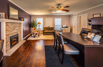 Clubroom/leasing office with wood flooring and two-tone paint.  There is a fireplace to the left of the room and photo. In the front of the photo to the right is a brown desk and two chairs.  Past the desk and fireplace is a chair and loveseat. - Photo Gallery 7
