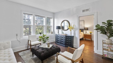 A living room with a black ottoman, white chair, and a large window.