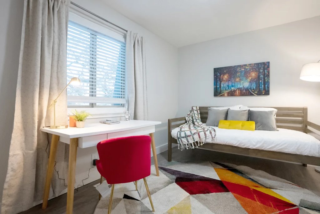 A room with a red chair, a white desk, and a white bed with a colorful rug.