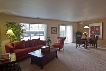 Living/Dining Area Living area of apartment with large picture window and doorwall to right along back wall. Red couch with lamp and tree behind, dark wood coffee table in front and red chair to right. Dining table with chairs in background with artwork on wall. Two-tone paint with white trim and beige carpet. - Photo Gallery 2