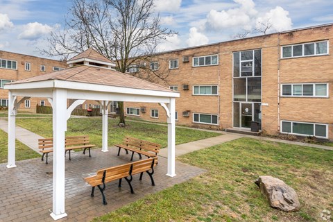 A white pavilion with benches is in front of a brick building.