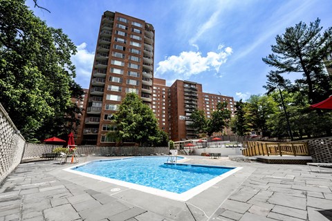 A swimming pool surrounded by a stone patio and trees with a red umbrella and chairs.