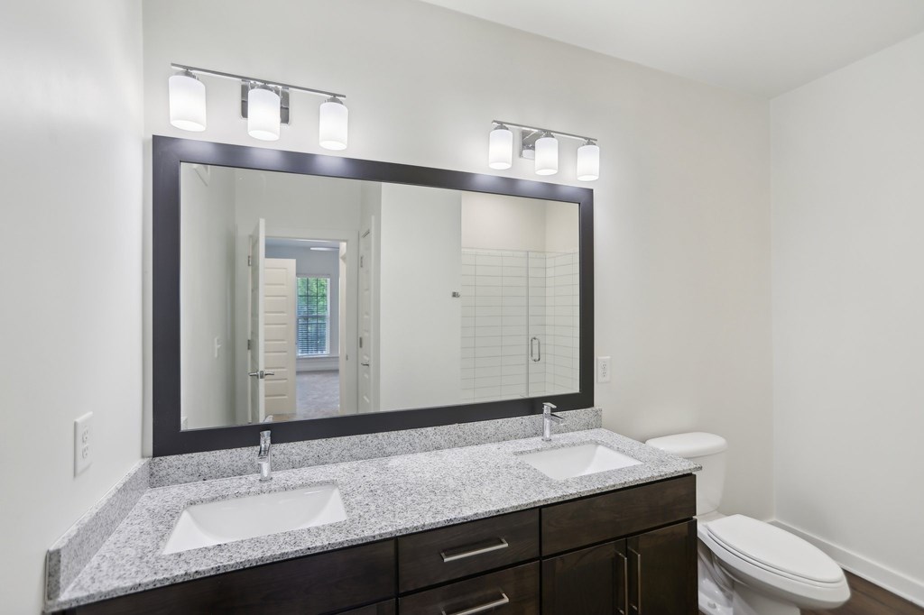 A bathroom with granite countertops, two sink, mirror, stand up glass panel shower, and toilet.